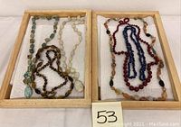 Two wooden trays each holding three mineral stone necklaces on white padding. Varied colors and bead shapes including blue-green, white, tiger eye brown, red, and multicolor agate beads.