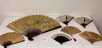 Full view of all seven oriental hand fans arranged flat showing various designs and sizes.