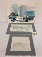 Photo shows three framed artworks: one watercolor painting of sailboats propped behind two pencil drawings framed with grey matting.