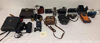 Collection of vintage cameras and binoculars displayed on table, showing various brands and models with cases and straps.