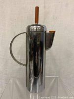 Front angle view of tall cylindrical chrome cocktail shaker with curved handle, spout with bakelite cap, and bakelite finial on lid.