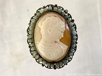Front view showing the carved white cameo of a saint set on brown background framed by greenish beads and metal filigree