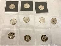Ten uncirculated nickels arranged in two rows; three nickel coins housed in black coin holders on the top row and seven nickels in transparent sleeves on the lower row, coins are bright and show no signs of wear.