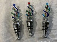 Three metal-based wine bottle stoppers with colorful twisted blown glass tops in blue, red, and light blue balls on top, arranged on white fabric.