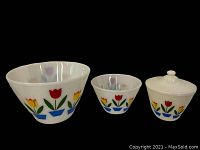 Set of three white milk glass Fire King casserole dishes with a tulip design in red, yellow, green, and blue. The dishes are arranged including the one with lid.