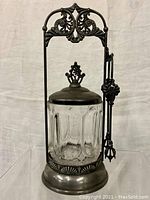 Front view of Victorian Art Nouveau pickle jar in silver plated framework with crystal jar and lid. Tongs hanging to the side.