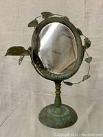 Front view of an oval brass vanity mirror with a green patina and decorative brass leaves and vines around the frame, mounted on a pedestal base.