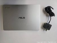 Closed top view of silver ASUS Chromebook laptop with charger cable and power adapter laid beside it.