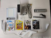 Complete set of Nintendo Wii console in white with vertical stand, cables, remote, Nunchuk, Wii Wheel, and four Wii game cases displayed on a white surface.