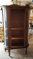 Frontal view showing wooden china cabinet with missing front glass and curved glass sides. Cabinet has three interior shelves and stands on four cabriole legs with small wheels.