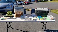 Photo showing full lot arranged on white folding table outdoors. Items include fishing basket, coolers, fishing net, tackle containers, bail bucket, and decor fish.