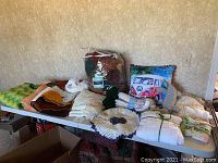 Overview of assorted blankets, afghans, doilies, and decorative pillows including Volkswagen print pillows and queen-size comforter spread on table.