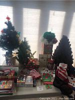 Photo showing two smaller decorated artificial Christmas trees, stockings, Christmas cards, and assorted ornaments, as well as some boxed ornaments and holiday decor items.