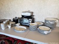 Wide shot displaying stacked dinner plates, smaller plates, bowls, cups with saucers, and small appliances on a table.
