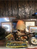 Wide view showing lamp with carved ram base surrounded by framed prints of ducks, bald eagle, golden lab, and needlepoint artwork, brass picture lights, and various hunting collectibles.