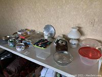 Photo showing kitchen items including cartoon mugs, a metal kettle, glass bowls, Rubbermaid containers, and a white hobnail lamp on a table.