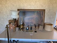 Framed print on a table with lamps, lampshades, photo frames, and figurines for sale.