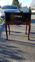 Front view of the small wooden table with Queen Ann legs, open top shelf with cable cut-out, photographed outdoors with a vehicle in background.
