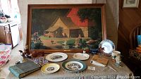 Photo of framed painting on table with plates, photo books, music box, and other items