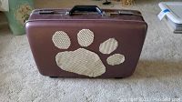 Brown hard-sided American Tourister suitcase with paw print fabric cutout on the side, placed on carpet.