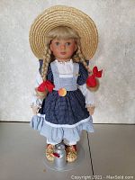 Front view of Margarete Steiff Babette doll in blue dress, white blouse, straw hat, red ribbons in braids, displayed on stand with metal milk can accessory.