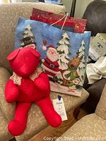 Photo of a large blue holiday gift bag featuring Santa Claus, reindeer, and trees with multiple red gift bags inside, seated on a chair with a red teddy bear in front.