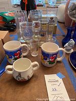 Photo of assorted German beer glasses, two Christmas ceramic mugs, and the Wedgwood Peter Rabbit children's mug with hedgehog cover on table