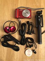 Red Lumix digital camera with multiple charging and audio cables, red Bluetooth earphones, earbuds, and black selfie stick on wooden surface.