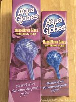 Boxes of Aqua Globes with purple packaging showing the product image and branding, highlighting hand-blown glass watering bulb and tagline.