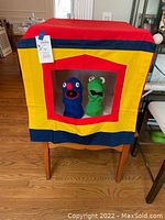 Front view of the colorful handmade puppet theater with the two crocheted puppets (green frog and blue monster) visible through the red window opening.