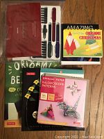 Calligraphy pen set with instruction booklet and markers, some origami instruction books including Christmas-themed, and various origami paper packs with colorful patterns and animal models on the covers.