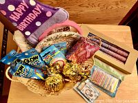 Basket with party items including birthday headband, decorative plates and napkins, glow bars, candle packs, and gold balloon holders.