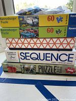 Photo of stacked boxes showing two Ravensburger 60-piece puzzles featuring a train and cars, a Super 3D puzzle with wildlife image, Sequence board game, and a box labeled 'A Game of Strategy' possibly Backgammon.
