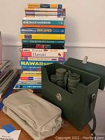 Stack of various travel books including guides for Paris, Provence, Riviera, Rome, Colorado, Ireland, Mexico, Hawaii, Europe, and retirement. Also shows the beige zippered pouch and the green vintage Selsi binoculars in their case.