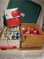Photo showing the large green wreath box with lid, new packaged red artificial tree storage bag, and two open boxes with assorted vintage glass ornaments of various colors including blue, white, and gold.