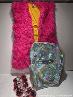 Photo showing hot pink fuzzy light-up vest hanging over yellow garment, rainbow iridescent LEGO backpack with two raised LEGO brick sections, and several pairs of red-frame fun sunglasses laid in front.
