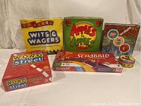 Seven family board games and one coding app game box arranged showing fronts with titles Wits & Wagers, Apples to Apples Junior, Bitsbox, Word on the Street (2 versions), Scrabble Crosswords game and card tin.