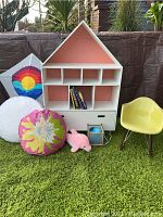 Photo of the white house-shaped bookcase with four cubby shelves, a bottom drawer, coral pink interior, surrounded by throw pillows, plush toy, child’s rocking chair on green rug.