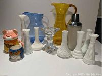 Photo of 7 vintage milk glass vases, two glass pitchers (yellow and blue), crystal candle holder, metal cocktail shaker, and two ceramic animal-themed cups with lids arranged in a row against white background.