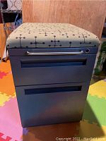 Front view of rolling black metal office bench with padded beige and blue patterned seat and two black drawers