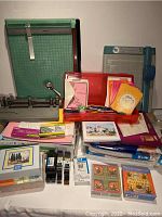Photo showing various office cutting tools including paper trimmers and industrial hole punch, also stationery cards, pencils, and pens in an orange container along with packages of envelopes and paper.