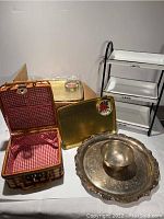 Photo shows child picnic basket open with red and white checkered fabric interior, gold trays, silver-plated dip platter and bowl, and white metal 3-tier tabletop shelf.