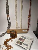 Four macrame plant holders hanging on a white backdrop, three unplanted holders and one holding a ceramic pot with drain hole. The colors of the holders are gray, cream (two), and reddish-brown with beads. The ceramic pot is brownish ceramic with a glossy glaze and is suspended by an orange macrame holder.