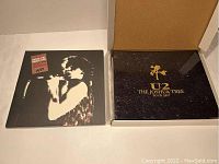 Collector's box with black leather-like cover embossed in gold reading 'U2 THE JOSHUA TREE TOUR 2017' and a smaller U2 vinyl record or booklet next to it.
