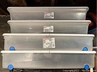 Four Sterilite plastic under bed storage containers stacked, showing translucent bodies with white lids and blue wheels.