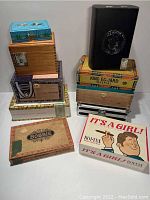 Photo showing a stacked assortment of 14 cigar boxes, both vintage and modern, various brands including King Edward Imperial, Bobbie, Titan Maduro, and novelty 'It's a Girl!'
