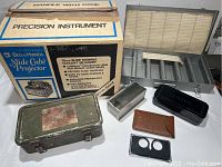Photo of Bell & Howell Slide Cube Projector box, metal military first aid kit, plastic slide organizers, and small containers