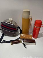 Overview showing the yellow thermos, red scout flask, felt-covered canteen, Tasco pocket telescope with box and case, and a small book.