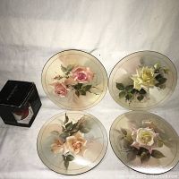 Four Keirstead hot plates arranged in a square showing various hand-painted rose designs in pastel colors, with a boxed mug placed next to them on a white cloth background.