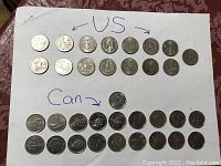 Top-down photo showing 15 US quarters arranged in two rows at the top and 19 Canadian quarters arranged in three rows at the bottom on a white sheet of paper with handwritten labels 'US' and 'Can'.